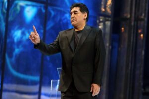 Diego Armando Maradona in abito elegante con il dito alzato su sfondo blu
